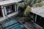 From above of wooden cottage with balcony located among tropical exotic trees with blue pool
